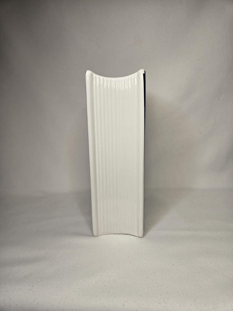 May include: A white ceramic object with a vertical ribbed texture and a concave top and bottom edge. The object has a rectangular profile with rounded corners and a smooth, glossy finish, standing upright on its side to display the ribbed surface.