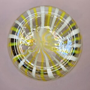 May include: Clear glass decorative bowl with a striped pattern in yellow, white, and black. The bowl has a rounded shape and a swirling design in the center. The text "by ANTHRO" is visible on the side.