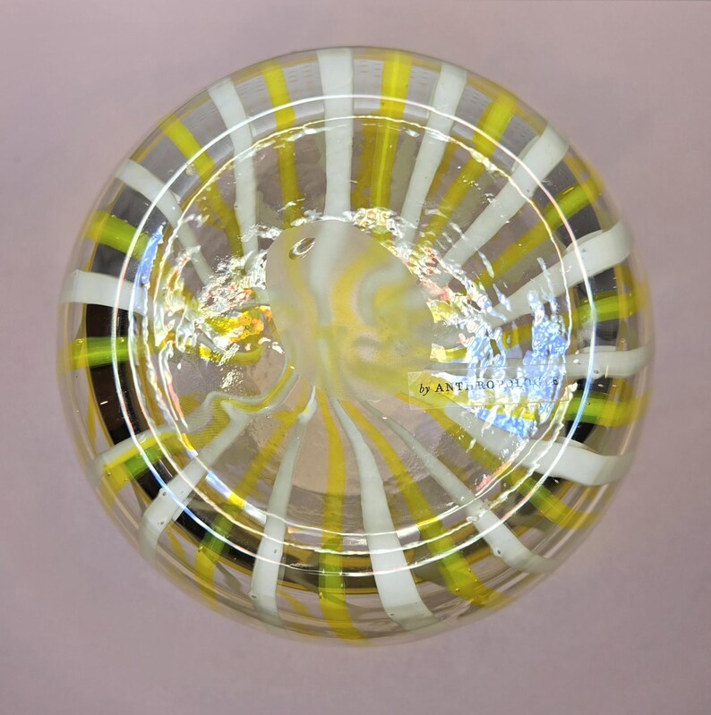 May include: Clear glass decorative bowl with a striped pattern in yellow, white, and black. The bowl has a rounded shape and a swirling design in the center. The text "by ANTHRO" is visible on the side.
