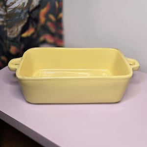 May include: A rectangular, light yellow ceramic baking dish with rounded corners and two small handles. The dish is empty and sits on a light purple surface. The background is a neutral color.