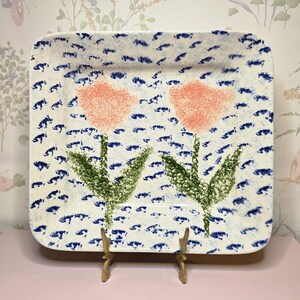 May include: Square ceramic serving platter with a white background and a blue dotted pattern. The platter features two pink tulip flowers with green stems and leaves. The platter is displayed on a gold-colored stand.