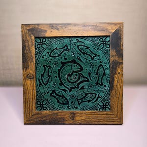 May include: Square decorative tile with a teal fish design in a wooden frame. The teal tile features a central fish surrounded by smaller fish and floral patterns. The wooden frame has a rustic, brown finish.