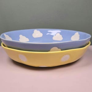 May include: Two ceramic bowls, one stacked on the other. The top bowl is light blue with white cloud designs. The bottom bowl is yellow with white sunburst designs. The bowls are on a pink surface.