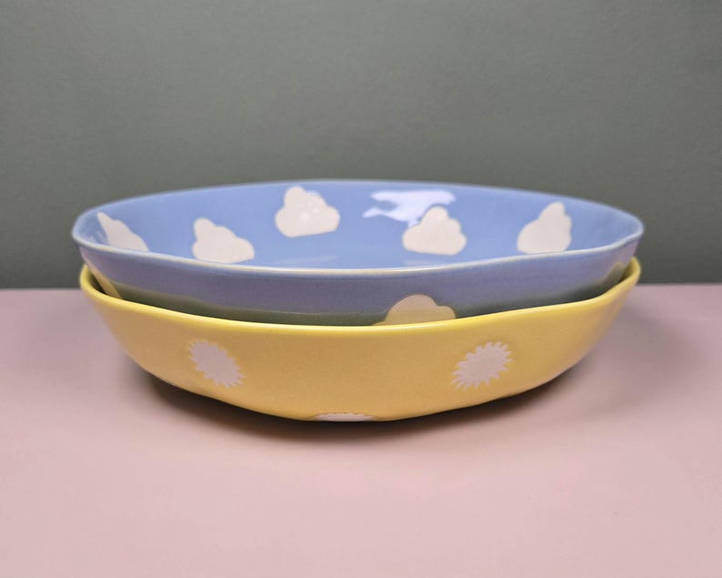 May include: Two ceramic bowls, one stacked on the other. The top bowl is light blue with white cloud designs. The bottom bowl is yellow with white sunburst designs. The bowls are on a pink surface.