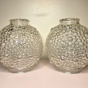 May include: Two clear glass light shades with a textured, bubble-like surface. The shades are round with a slightly flared base and a textured rim. They are likely for a ceiling light fixture.