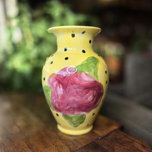 May include: A yellow ceramic vase with a hand-painted pink rose and green leaves. The vase has black polka dots and a flared rim. The vase is sitting on a wooden surface.