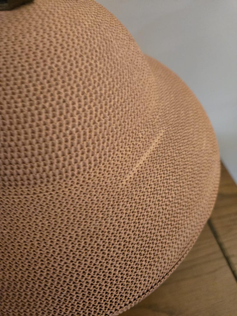 May include: Close-up of a tan hat featuring a textured, woven design. The hat's surface has a repeating geometric pattern, giving it a tactile feel. The hat's shape is rounded, indicating a casual style.