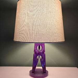 May include: A unique table lamp with a purple clothespin-shaped base and a beige lampshade. The base has a circular bottom and two circular cutouts. The lampshade is a light tan color with a dark blue trim.