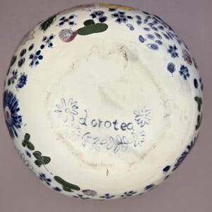 May include: A white ceramic bowl with a floral design in blue, green, yellow, and red. The bottom of the bowl has the name "Dorotea" written in blue script, surrounded by floral and leaf motifs.