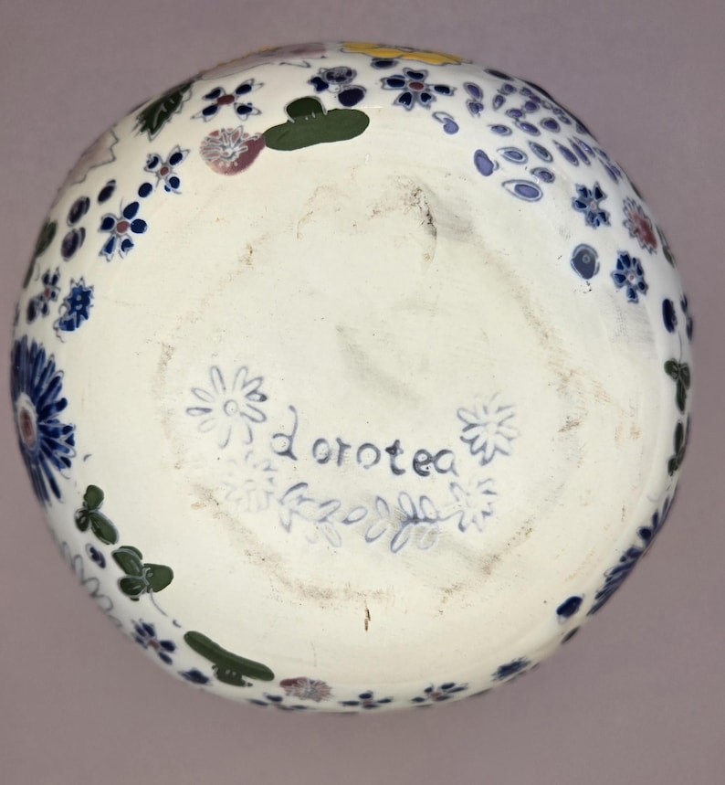 May include: A white ceramic bowl with a floral design in blue, green, yellow, and red. The bottom of the bowl has the name "Dorotea" written in blue script, surrounded by floral and leaf motifs.