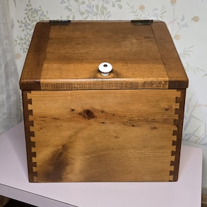 May include: A wooden box with a hinged lid and a white knob. The box is constructed with dovetail joints and has a natural wood grain finish. The lid is slightly angled, and the box appears to be a storage container.