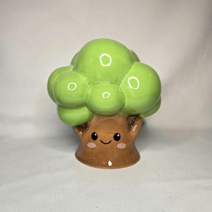 May include: A ceramic tree-shaped figurine with a smiling face. The tree has a brown trunk and a bright green, rounded top. The figurine is a decorative item, likely for home decor.