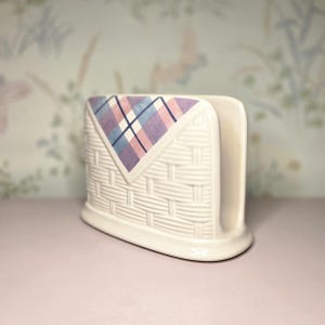 May include: A white ceramic napkin holder with a woven basket design. The top features a decorative plaid pattern in shades of pink, blue, and purple. The holder is designed to hold napkins.