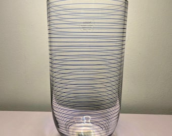 May include: A clear glass cylindrical vase featuring thin, horizontal blue lines wrapping around the exterior. The glass surface displays a faint, circular brand logo near the top and a small rectangular sticker visible at the base.