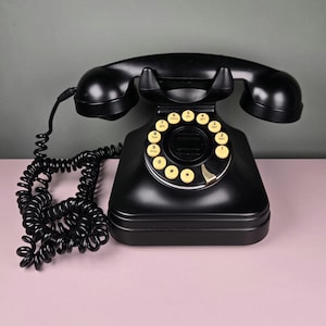 May include: A vintage black rotary dial telephone with a handset and coiled cord. The phone has a circular dial with yellow number buttons and a black base. The phone is on a pink surface.