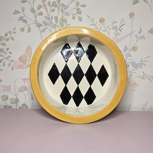 May include: A round ceramic bowl with a yellow rim and a cream-colored interior. The interior features a black and white diamond pattern. The bowl is set against a floral wallpaper background.
