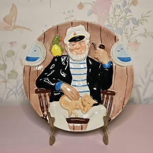 May include: A decorative ceramic plate featuring a seated sailor with a white beard, captain's hat, and pipe. The sailor is wearing a blue and white striped shirt and black jacket. A sleeping orange cat rests on his lap, and a green parrot sits on his shoulder.