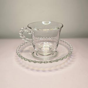 May include: A clear glass teacup and saucer set. The teacup has a beaded handle and a beaded rim. The saucer also has a beaded edge. The set is made of clear glass.
