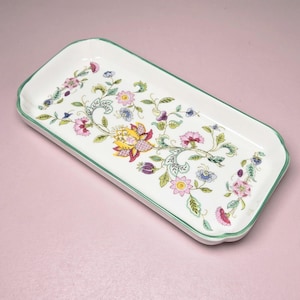 May include: A rectangular white ceramic tray with rounded corners and a green rim. The surface features a colorful floral pattern with pink, yellow, and blue flowers, green leaves, and winding vines arranged in a central decorative design.