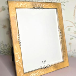 May include: A rectangular picture frame with a light yellow marbled finish and a silver beaded trim. The frame has a white interior and is labeled "8.10". The frame is propped up on a black stand.