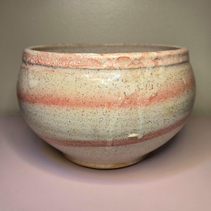 May include: A round ceramic bowl with a speckled cream-colored glaze featuring horizontal bands of soft pink. The rim has a textured, uneven finish with visible drips of glaze running down the sides of the vessel, creating a layered appearance.