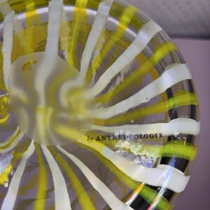 May include: A clear glass bowl with a spiral design of yellow and white stripes. The bowl has a frosted center and the text "by ANTHROPOLOGIE" is visible. The bowl is a decorative item.