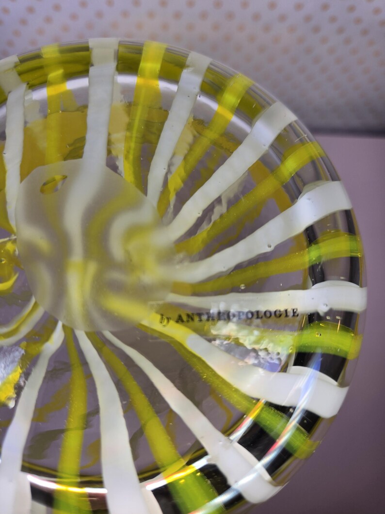 May include: A clear glass bowl with a spiral design of yellow and white stripes. The bowl has a frosted center and the text "by ANTHROPOLOGIE" is visible. The bowl is a decorative item.