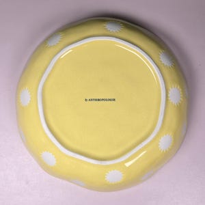 May include: A yellow ceramic bowl with a white interior rim and white starburst designs around the edge. The bowl has the text "by ANTHROPOLOGIE" printed on the interior.
