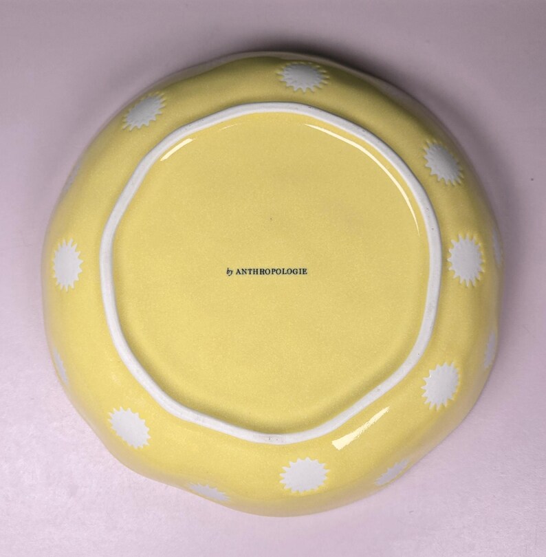 May include: A yellow ceramic bowl with a white interior rim and white starburst designs around the edge. The bowl has the text "by ANTHROPOLOGIE" printed on the interior.