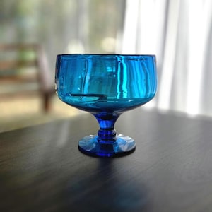 May include: A vibrant blue glass goblet with a rounded bowl and a short stem. The glass has a glossy finish, reflecting light. The goblet sits on a dark surface, with a blurred background.