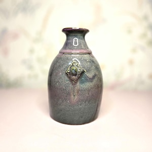 May include: A handcrafted ceramic vase with a gray and purple glaze. The vase features a decorative diamond-shaped embellishment and a narrow neck. The vase is approximately 7 inches tall, perfect for displaying flowers or as a decorative piece.