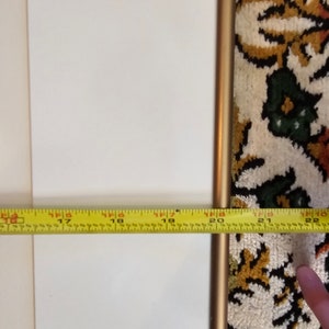 May include: A close-up of a white picture frame with a gold metal frame. A yellow measuring tape is placed at the bottom of the frame, showing a measurement of 45.7 cm.