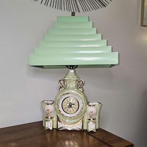 May include: A vintage table lamp with a mint green stepped shade. The base is a ceramic clock with a round face, gold accents, and floral details. Two small vases flank the clock.