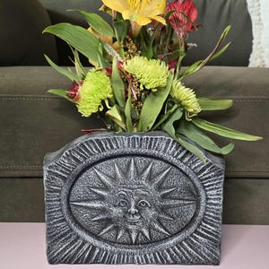 May include: A gray stone vase with a sun face design, holding a bouquet of yellow, green, and red flowers.