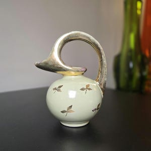 May include: A small, cream-colored ceramic creamer featuring a rounded body decorated with a repeating pattern of metallic gold leaf motifs and a prominent, arched metallic gold handle that extends from the base to the spout.