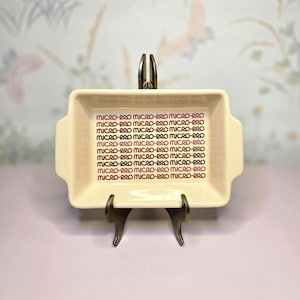 May include: A rectangular, off-white ceramic dish with the text "MICRO-PRO" in a repeating pattern in purple. The dish has handles on each side and is displayed on a decorative metal stand.