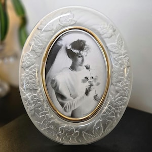 May include: An oval glass picture frame featuring a frosted finish with embossed floral and leaf patterns. The frame has a smooth gold-toned inner rim and contains a black and white photograph of a person wearing a veil and holding a single rose.