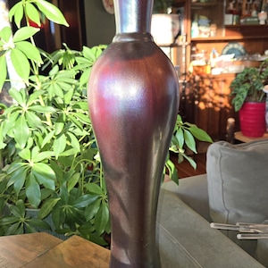 May include: A tall, iridescent vase with a slender neck and flared base. The vase has a dark, metallic finish with hints of purple and brown. It stands on a wooden surface, with green plants in the background. The vase is approximately 14 inches tall.