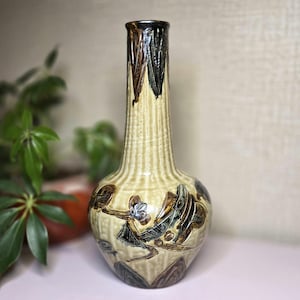 May include: A tall, ceramic vase with a narrow neck and a bulbous base. The vase is primarily beige with vertical ribbed detailing. It features brown and blue floral and abstract designs. The rim is dark brown and black.
