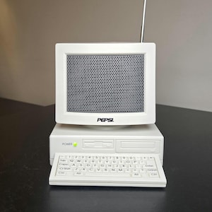 May include: A white novelty radio shaped like a vintage desktop computer with a monitor screen featuring a grey speaker grille and the Pepsi logo. The base includes a power button with a green light, a floppy disk drive slot, and a QWERTY keyboard.