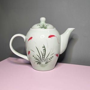 May include: A white ceramic teapot with a lid, handle, and spout. The teapot is decorated with red fish and aquatic plant designs in black and green. The teapot is sitting on a pink surface.