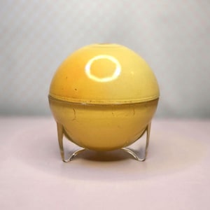 May include: A yellow, spherical container with a lid, resting on three clear, curved legs. The container has a slightly cracked surface and a glossy finish. The design is reminiscent of a vintage cosmetic or trinket box.
