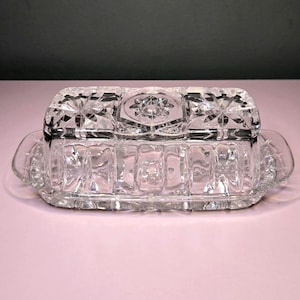 May include: A clear glass butter dish featuring a rectangular base with integrated handles and a matching lid. The lid is decorated with a geometric starburst pattern and a central hexagonal motif, while the base displays vertical panels.