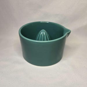 May include: A teal ceramic citrus juicer. The juicer has a conical reamer in the center and a pouring spout. The exterior is a solid teal color. The juicer is designed for easy citrus fruit juice extraction.