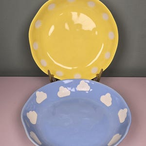 May include: Two ceramic plates. The top plate is yellow with white starburst designs around the rim. The bottom plate is light blue with white cloud designs around the rim. Both plates have a wavy edge.