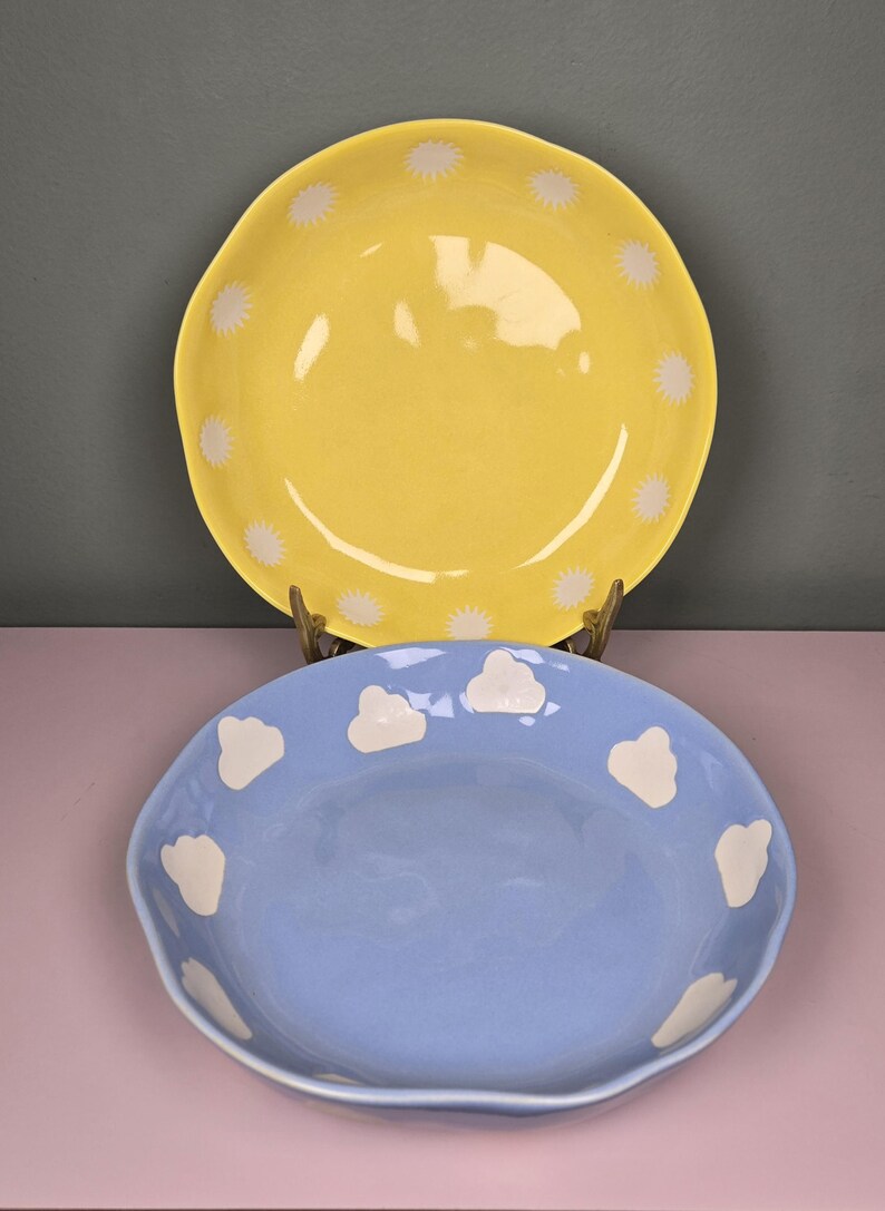 May include: Two ceramic plates. The top plate is yellow with white starburst designs around the rim. The bottom plate is light blue with white cloud designs around the rim. Both plates have a wavy edge.