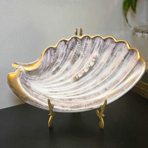 May include: A shell-shaped dish featuring a white and brown distressed finish with gold-painted scalloped edges. The interior surface has a ribbed texture and a small, central decorative element. The dish is supported by a gold-toned metal stand.