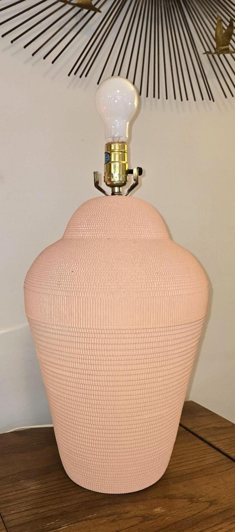 May include: A peach-coloured table lamp with a textured, ribbed surface. The lamp has a bulb and a brass-coloured base. The lamp is on a wooden surface.
