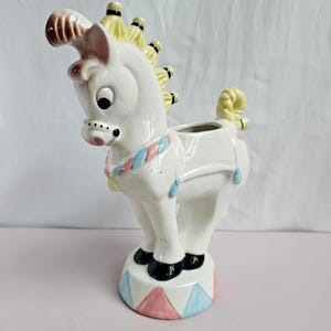 May include: White ceramic planter shaped like a horse with a yellow mane and tail. The horse is wearing a blue and pink striped collar and has a black and white face. The planter is standing on a pink and blue circus-themed base.