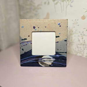 May include: Square ceramic picture frame with a neutral beige top and a dark blue and gray bottom. The frame has a white square opening in the center and is decorated with blue and purple paint splatters.